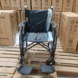 Standard manual wheelchair with safety brakes, foldable design, displayed on a wooden pallet among cardboard boxes.